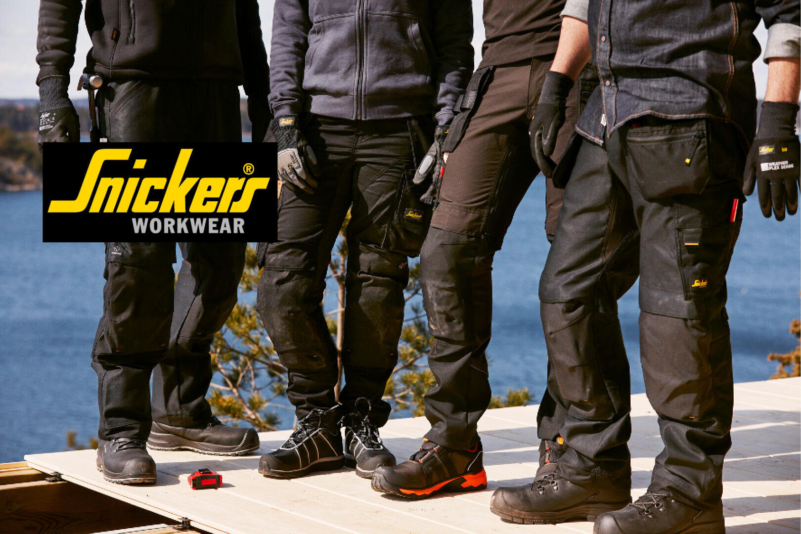 Quatre personnes vêtues de pantalons et de gants noirs Snickers Workwear se tiennent sur une plate-forme en bois en plein air au bord de l'eau, avec un logo Snickers Workwear dans le coin supérieur gauche. Seule la partie inférieure de leur corps est visible.