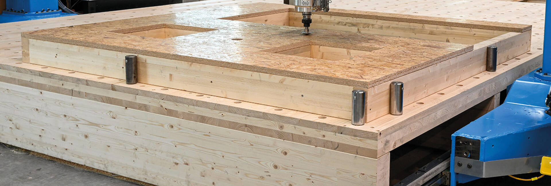 Eine CNC-Maschine schneidet Fensteröffnungen in eine große, vorgefertigte Holzwandplatte in einer Produktionsstätte.