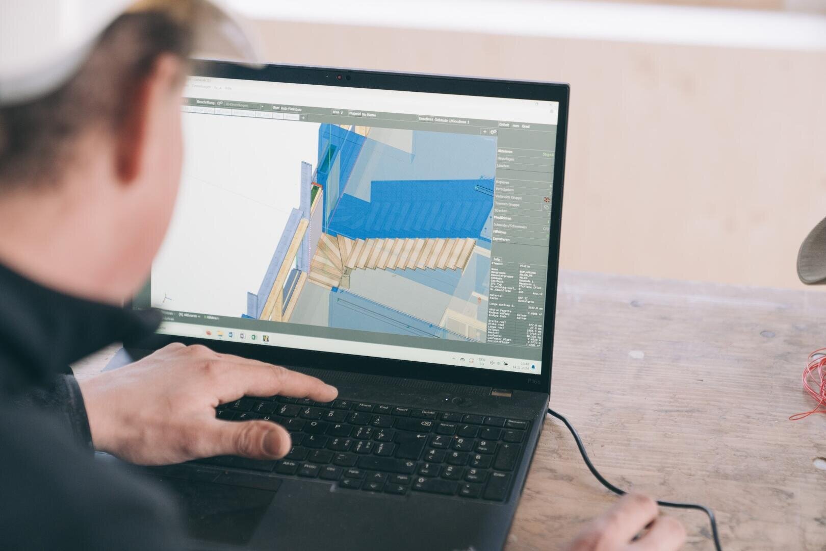 Eine Person arbeitet mit einem Laptop an einem 3D-Architektur- oder Ingenieursmodell mit blauen und beigen Strukturelementen auf einem Holztisch. Nur ihre Hände und ein Teil ihres Gesichts sind sichtbar.