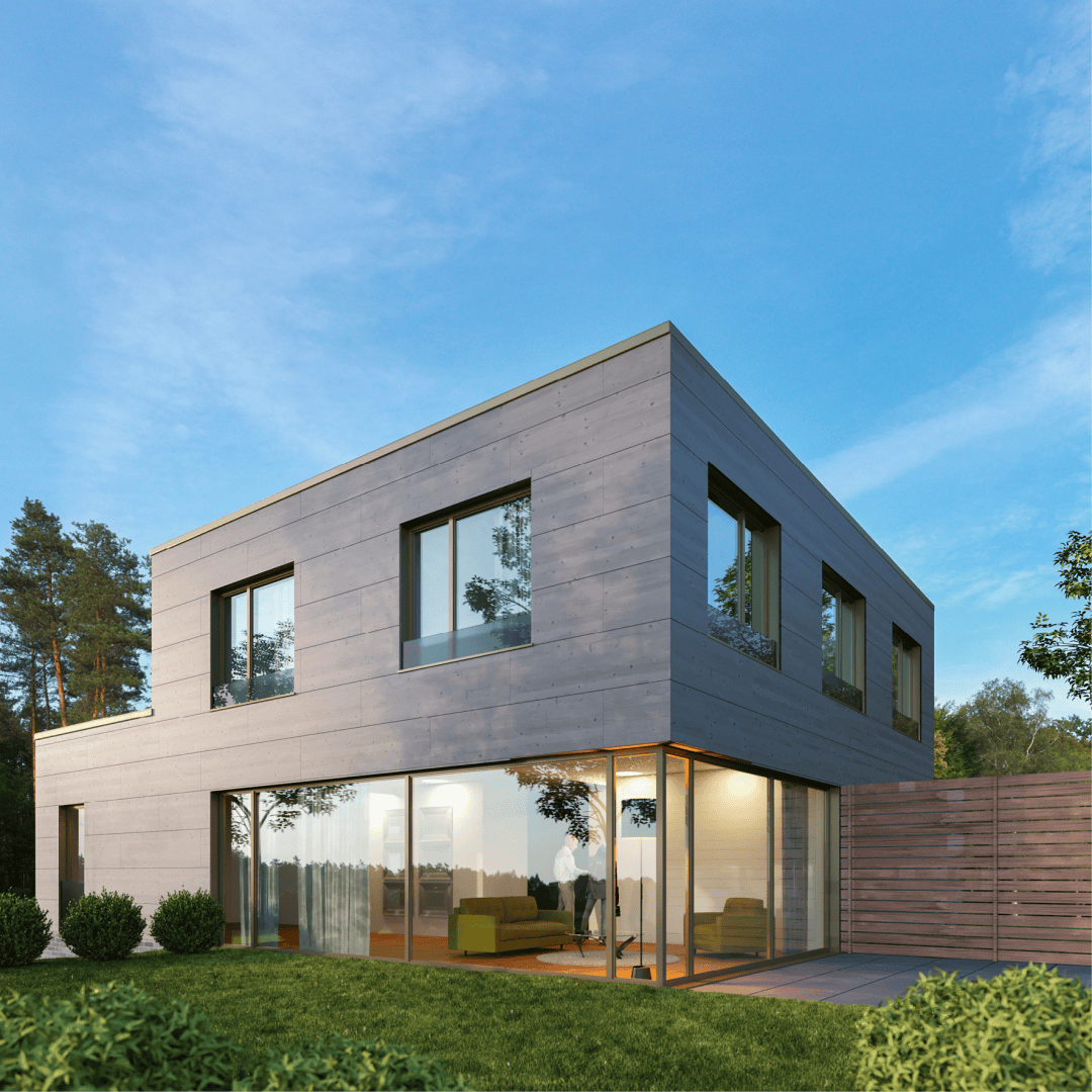 Maison moderne de deux étages avec de grandes fenêtres et des revêtements extérieurs gris, entourée d'espaces verts et d'une clôture en bois, sous un ciel bleu clair. A l'intérieur, une personne est visible à travers les portes vitrées.