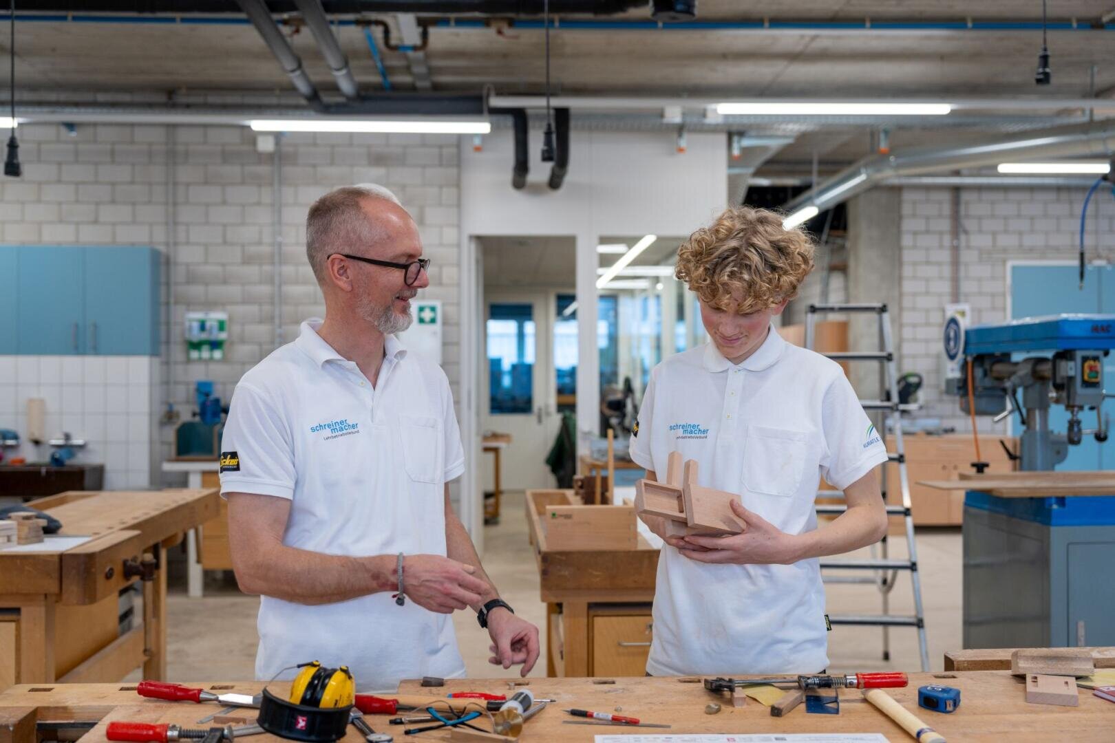 Deux personnes en polo blanc se tiennent dans un atelier. L'un d'eux, un homme âgé, sourit tout en discutant avec une personne plus jeune qui examine un projet en bois. En arrière-plan, on aperçoit des établis, des outils et du matériel pour travailler le bois.