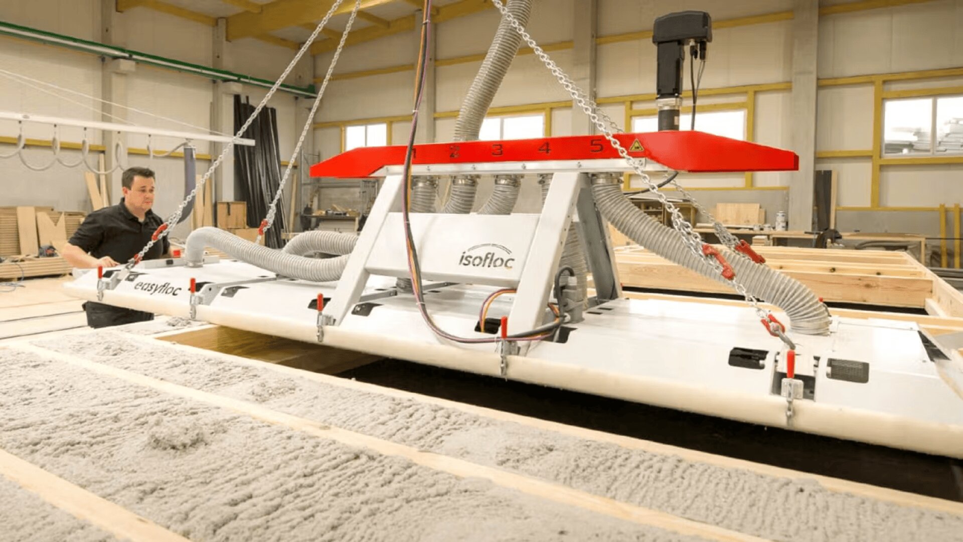 Un ouvrier utilise une grande machine d'isolation industrielle avec des tuyaux et un cadre rouge, répandant des matériaux d'isolation en vrac entre des poutres en bois dans un atelier.