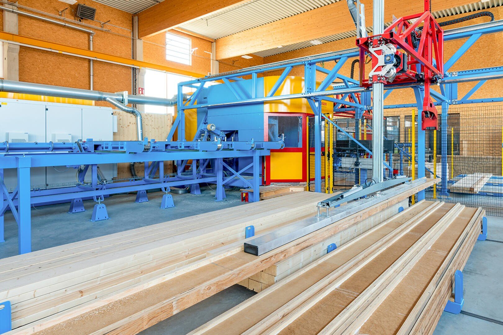 Holzbalkenstapel stehen in einer modernen Holzverarbeitungsfabrik mit blauen Metallmaschinen und automatisierten Anlagen unter einer hohen Industriedecke mit großen Fenstern.
