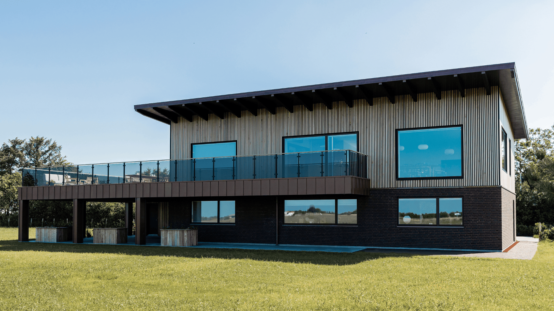 Une maison moderne à deux étages avec de grandes fenêtres teintées de bleu, des boiseries au niveau supérieur, des briques foncées au niveau inférieur et un balcon spacieux avec des balustrades en verre, entouré d'herbe verte et d'arbres.