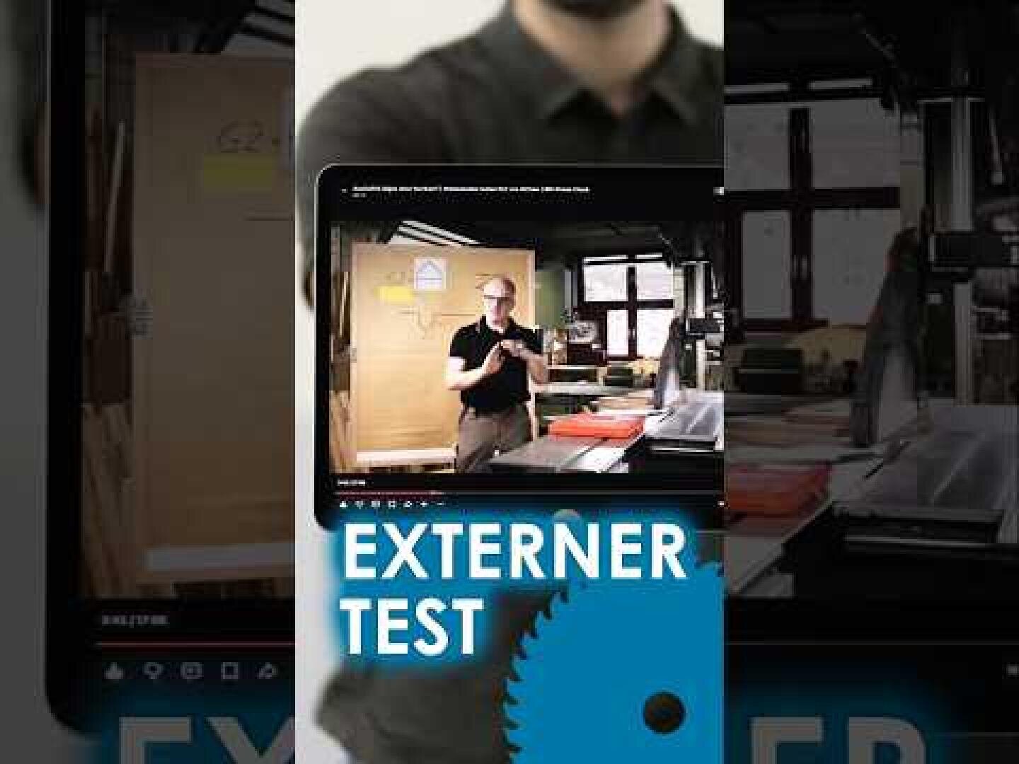Une personne tient une tablette sur laquelle est diffusée une vidéo montrant quelqu’un dans un atelier. L’écran affiche « EXTERNER TEST », avec une icône en forme de roue dentée bleue en dessous. Des outils et du matériel d’atelier sont visibles à l’arrière-plan.