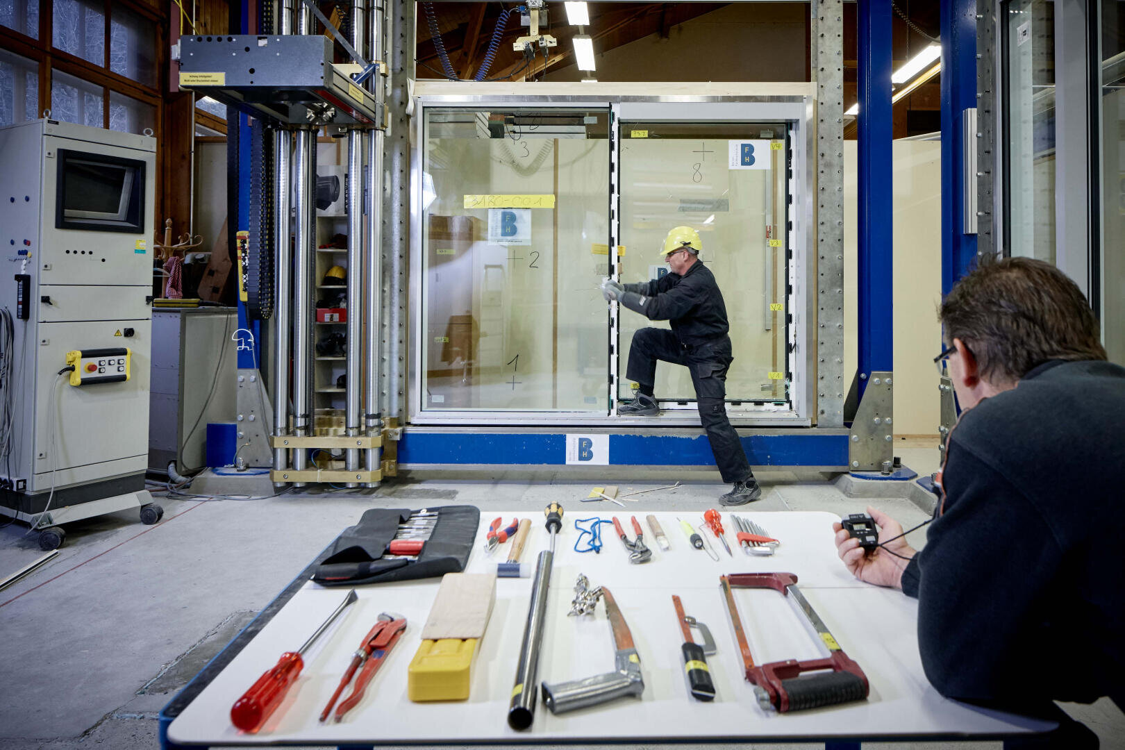 Eine Person in Schutzkleidung versucht, eine große Glastür in einer Laborumgebung aufzubrechen, während eine andere Person Daten beobachtet und aufzeichnet. Auf einem Tisch in der Nähe sind verschiedene Werkzeuge übersichtlich angeordnet.