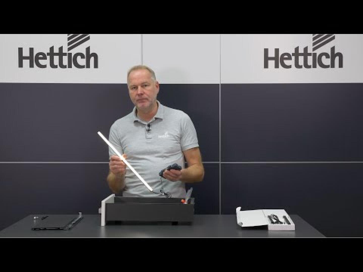 Un homme vêtu d'un polo gris clair se tient à une table et assemble un objet, tenant un outil d'une main et un composant noir de l'autre. Derrière lui, le mur affiche le logo Hettich. Des boîtes et des pièces sont posées sur la table.