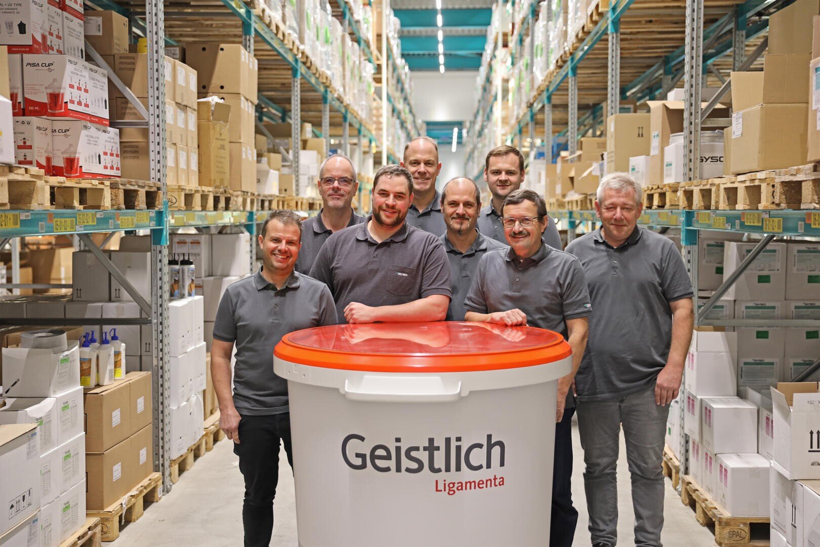 Acht Männer in passenden grauen Hemden stehen in einem Lagergang und lächeln hinter einem großen weißen Container mit der Aufschrift "Geistlich Ligamenta". Auf beiden Seiten des Gangs stapeln sich Regale mit Kisten und Zubehör.