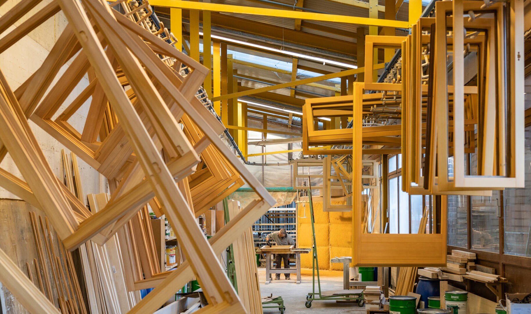 In einer hell erleuchteten Werkstatt hängen Holzrahmen an Regalen, und im Hintergrund ist ein Arbeiter zu sehen, der inmitten von Werkzeugen und Materialien an einem Tisch arbeitet oder etwas zusammenbaut. Überall im Raum sind gelbe Metallstrukturen und Holzbretter zu sehen.