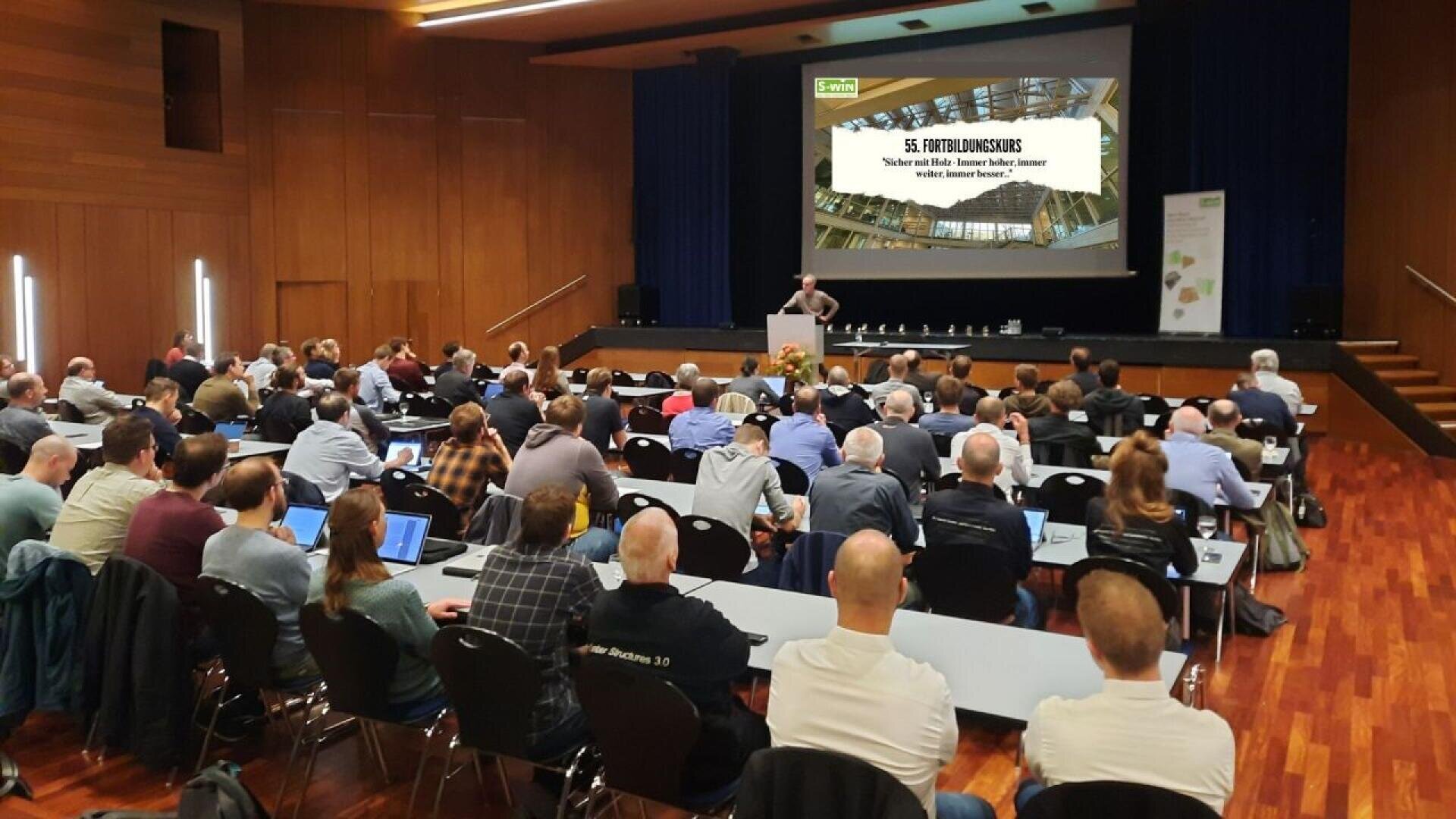 Ein Redner spricht auf der Bühne vor einem sitzenden Publikum in einem modernen Konferenzsaal. Auf einem großen Bildschirm hinter dem Redner wird der Titel eines Workshops zur 3D-Visualisierung angezeigt. Die Teilnehmer verwenden Laptops und machen sich Notizen.