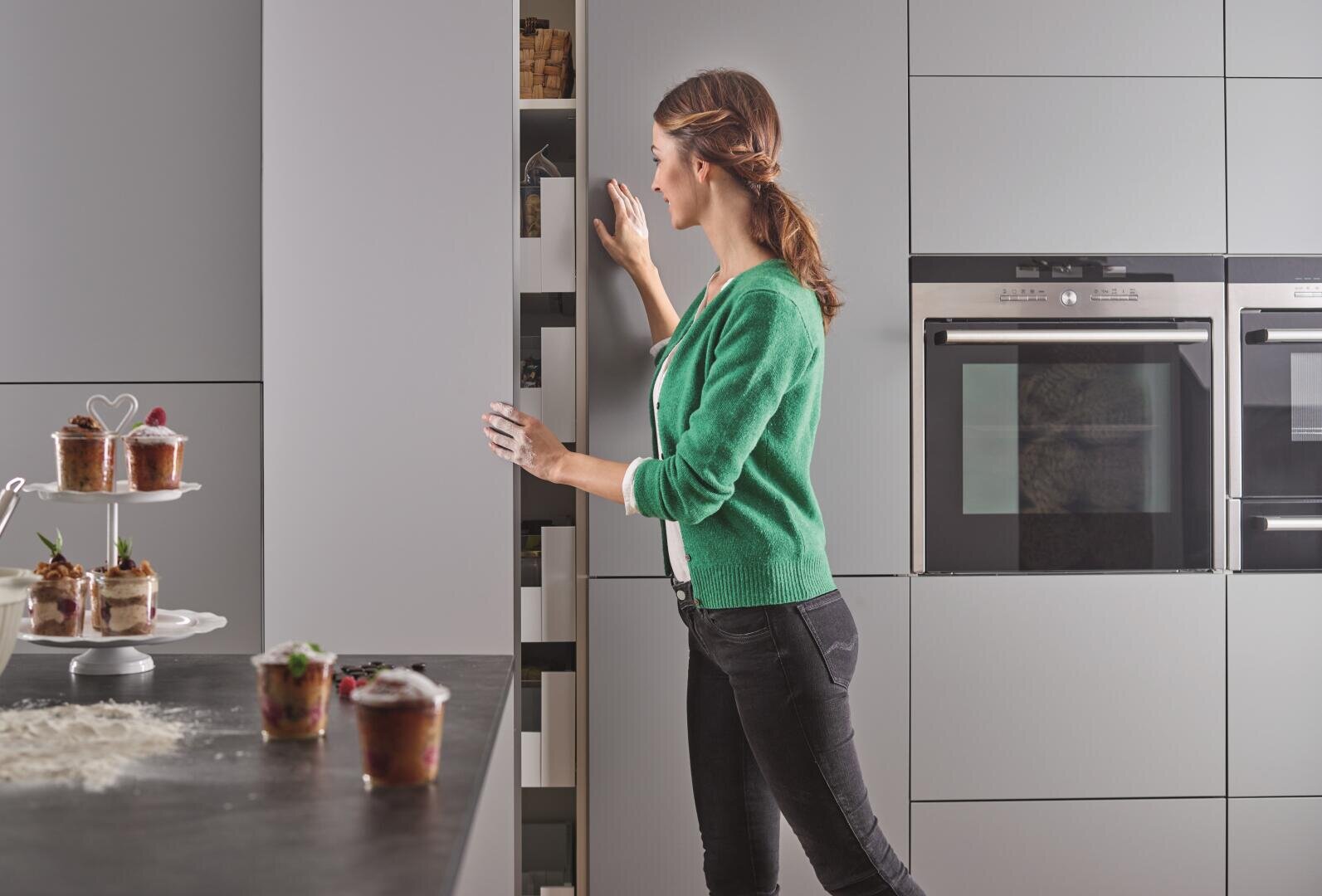 Eine Frau in einem grünen Pullover und schwarzen Jeans öffnet einen hohen, grauen Küchenschrank. Nebenan auf der Theke stehen Gläser und Desserts, im Hintergrund sind die eingebauten Öfen zu sehen.