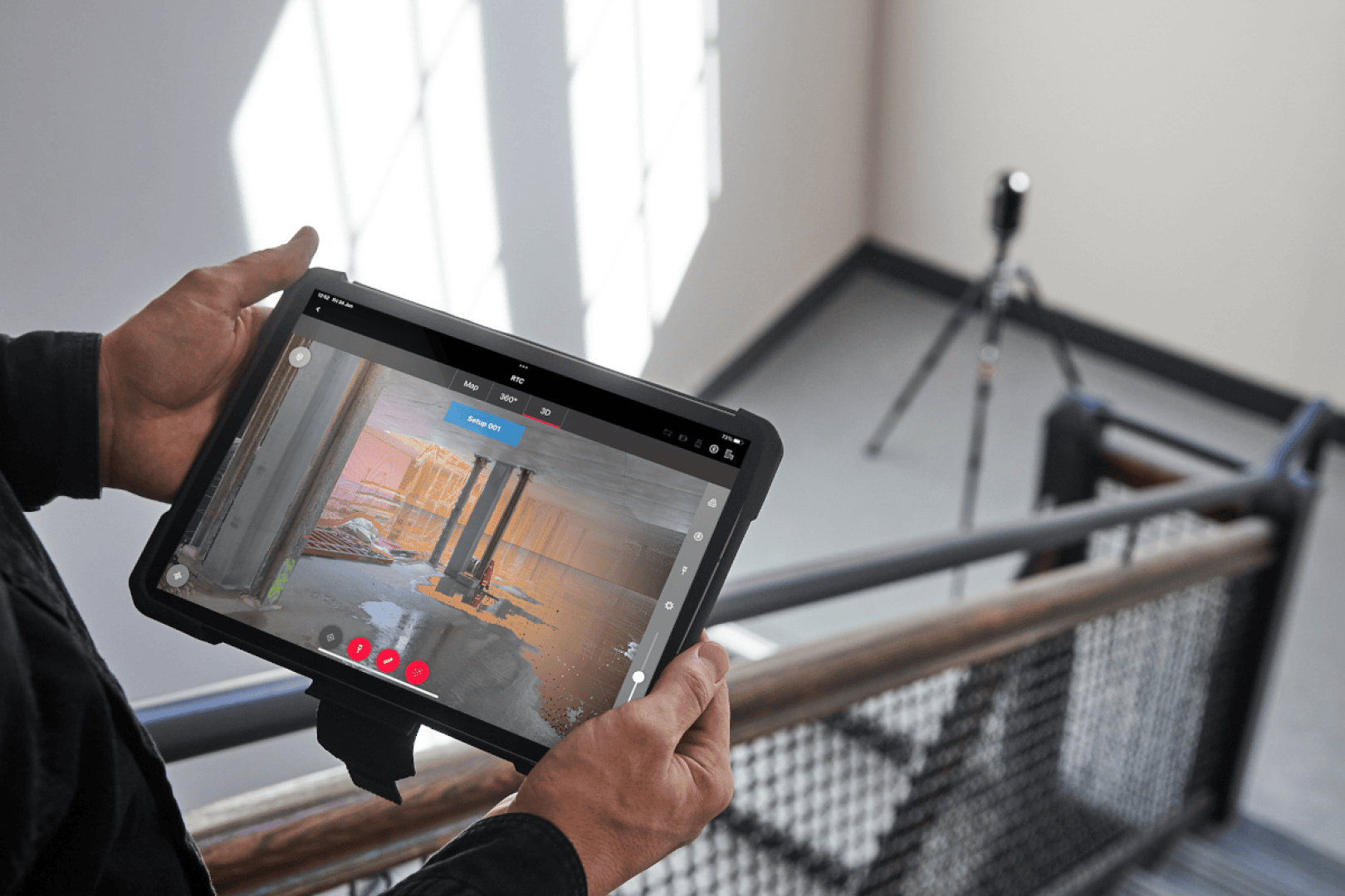 Eine Person hält ein Tablet, das einen 3D-Scan eines Raums zeigt, während ein auf einem Stativ montierter Scanner im Hintergrund am oberen Ende eines Treppenhauses Daten aufnimmt. Sonnenlicht fällt durch die Fenster auf den Boden und die Wände.