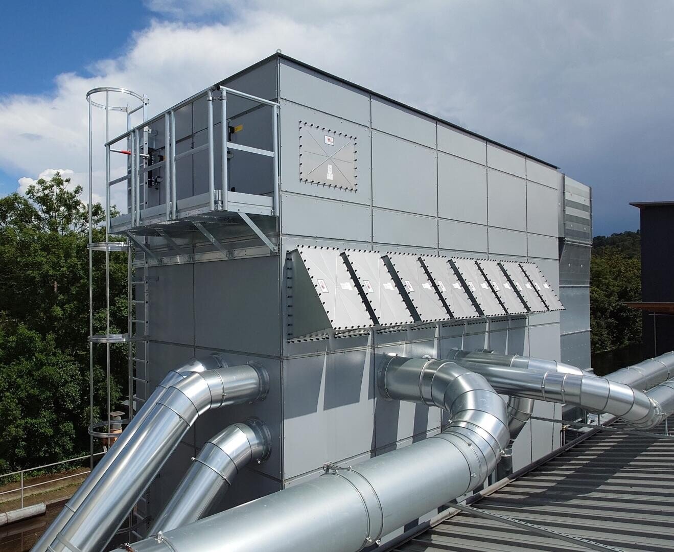 Eine große industrielle Luftfilteranlage mit mehreren Metallkanälen und Entlüftungsöffnungen ist im Freien neben Bäumen und unter einem teilweise bewölkten Himmel installiert. Eine Leiter führt zu einer Zugangsplattform an der Filteranlage.