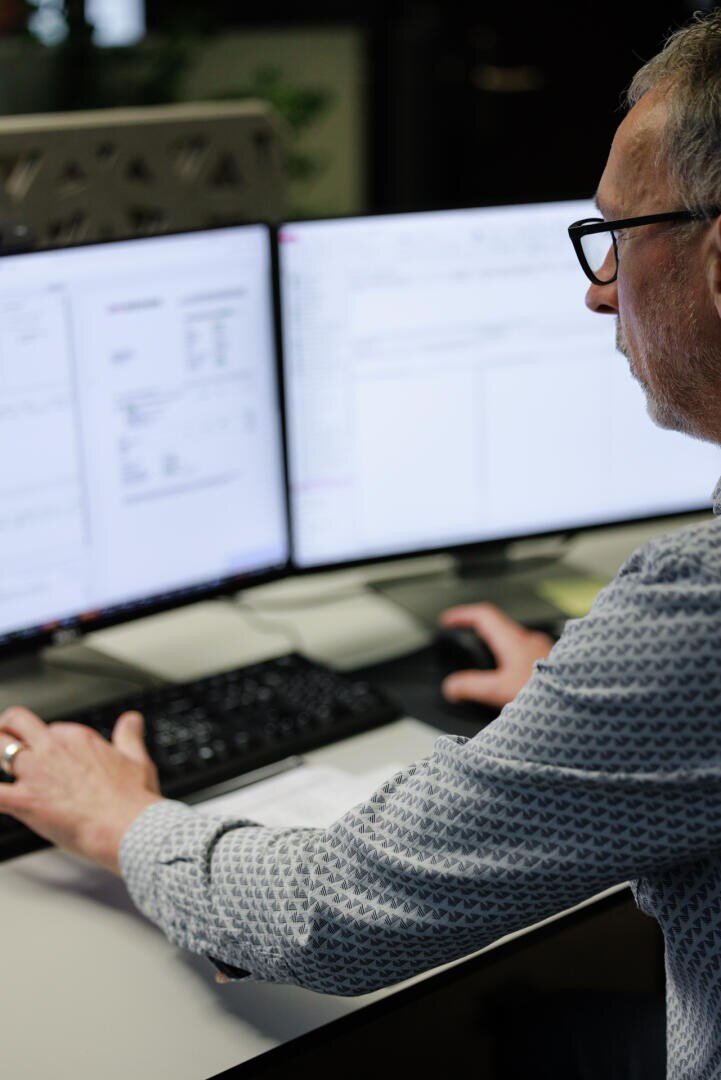 Ein Mann mit Brille arbeitet an einem Schreibtisch mit zwei Computermonitoren, auf denen Dokumente und Diagramme angezeigt werden, und benutzt eine Tastatur und eine Maus.