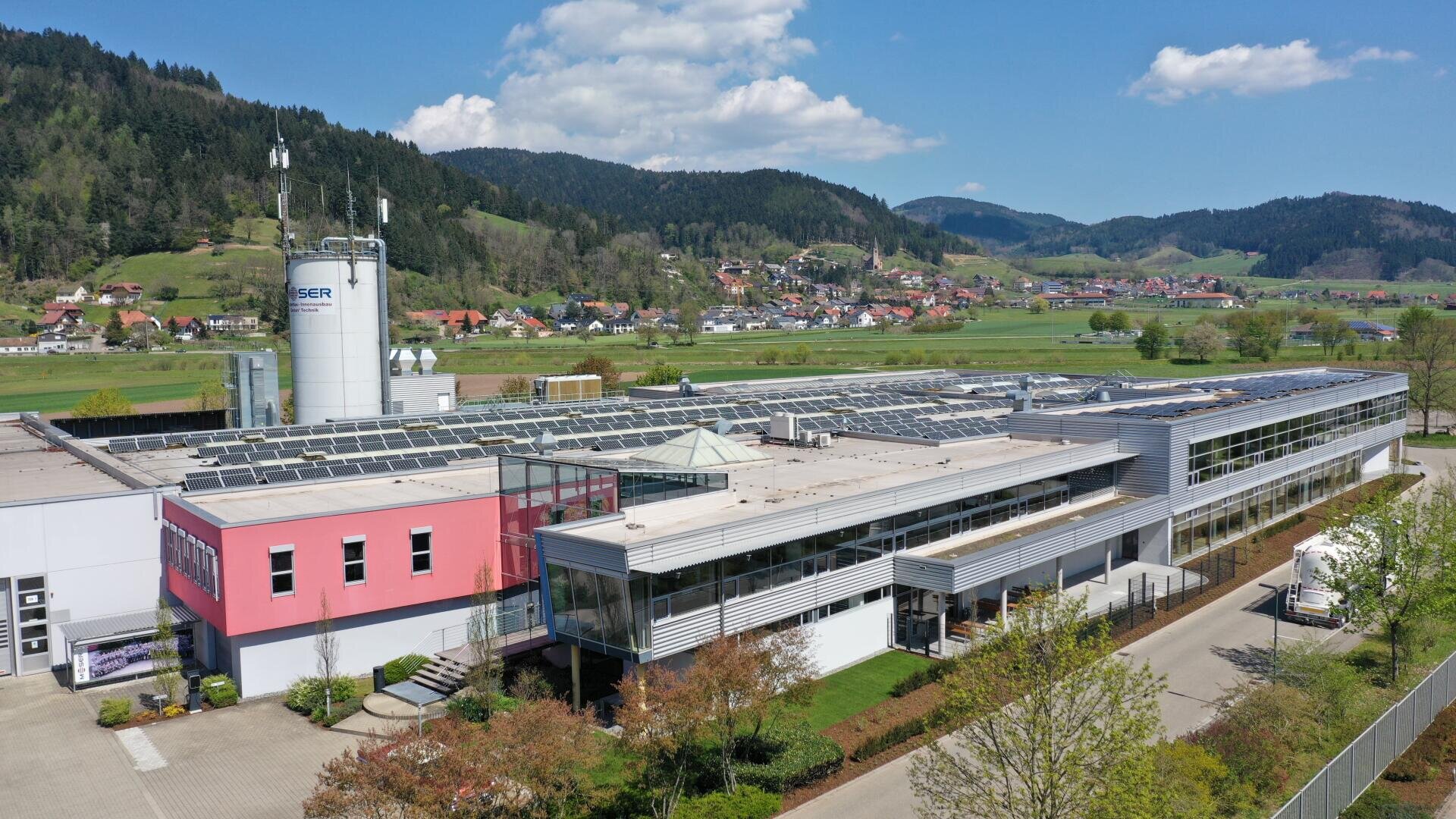 Ein modernes Industriegebäude mit Sonnenkollektoren auf dem Dach liegt in einem grünen Tal, umgeben von Bäumen und fernen Hügeln unter einem teilweise bewölkten Himmel. In der Nähe ist eine kleine Stadt inmitten der Landschaft zu sehen.