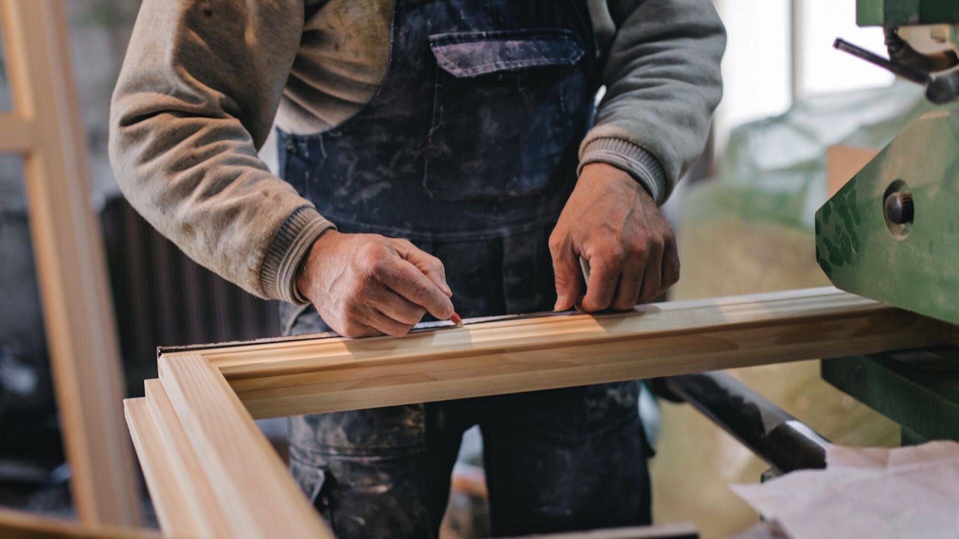 Eine Person im Arbeitsoverall misst und markiert einen Holzrahmen mit einem Bleistift in einer Werkstatt, um das Holz für den Zuschnitt oder die Montage vorzubereiten.