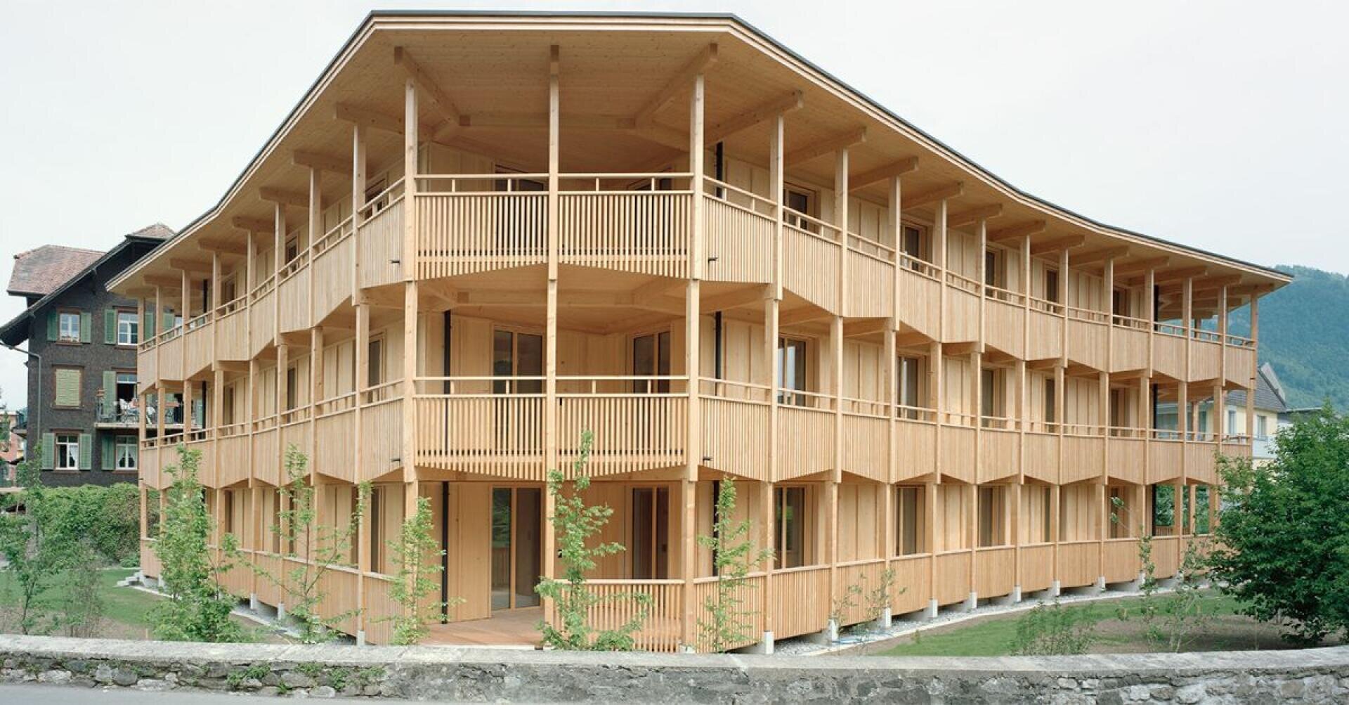 Ein modernes dreistöckiges Holzgebäude mit umhüllenden Balkonen und vertikalen Latten, umgeben von Grün und vor der Kulisse von Bäumen und Bergen.