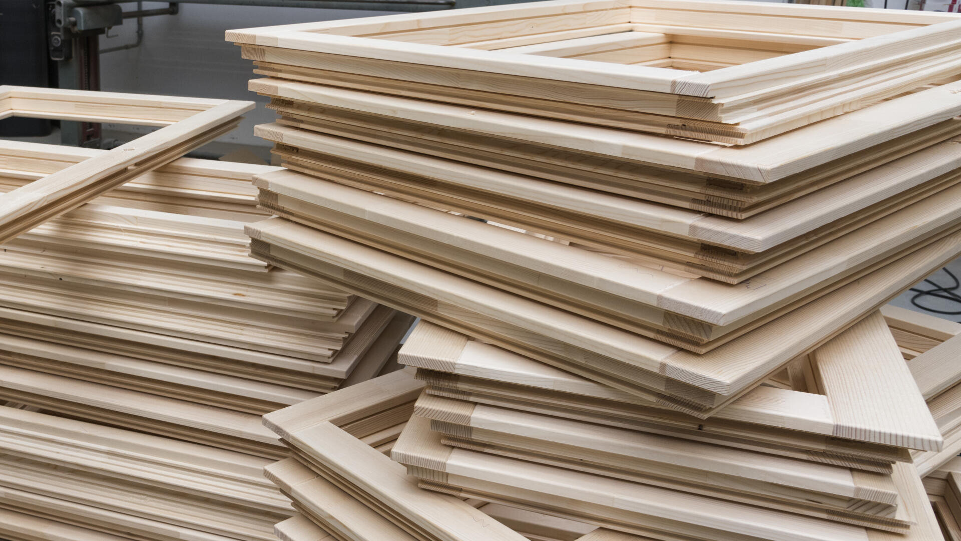 Dans un atelier, des piles de cadres en bois non finis sont empilées les unes sur les autres, prêtes à être transformées ou finies.