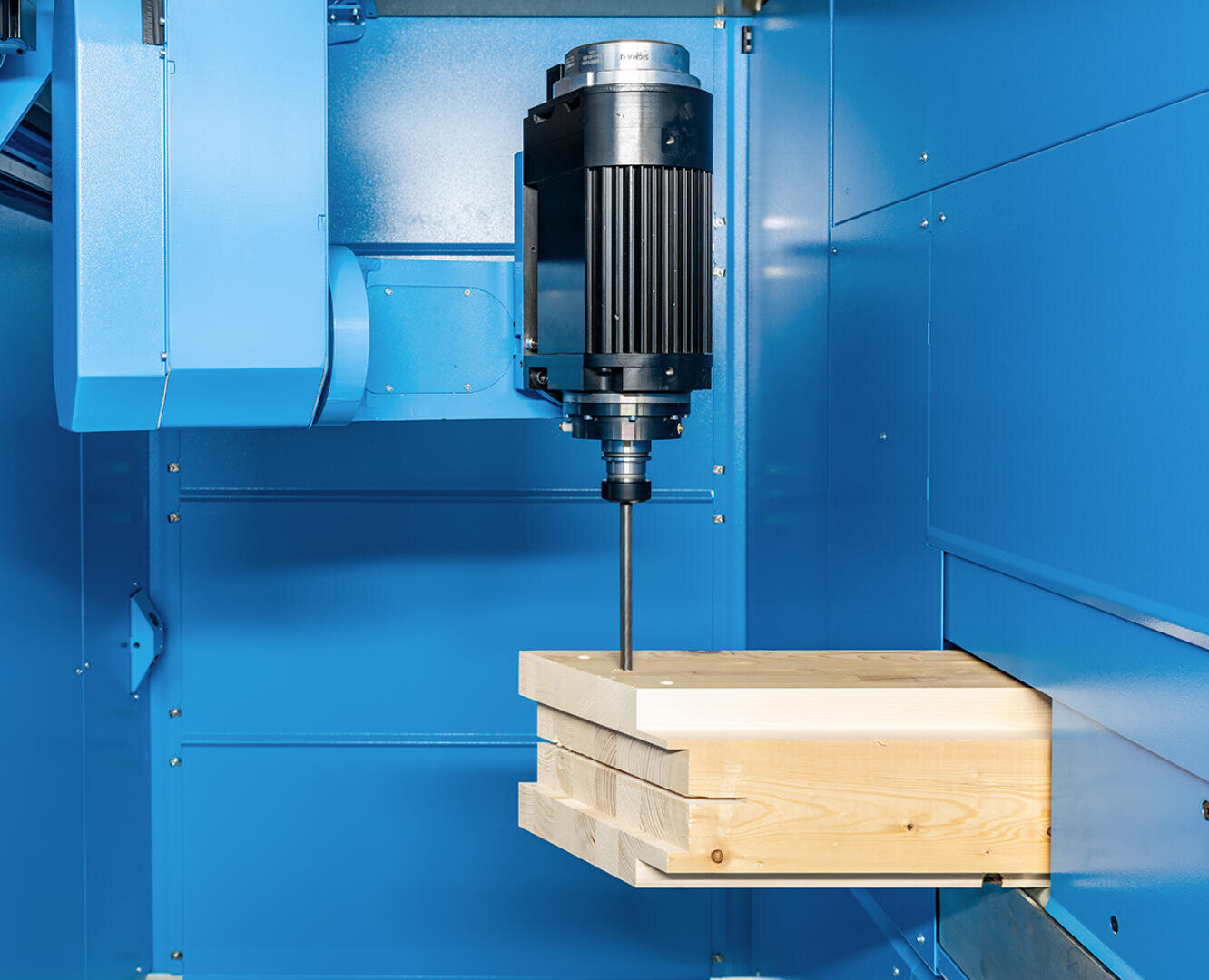 Eine CNC-Maschine mit vertikaler Spindel bohrt in gestapelte Holzplatten innerhalb eines blauen Gehäuses.