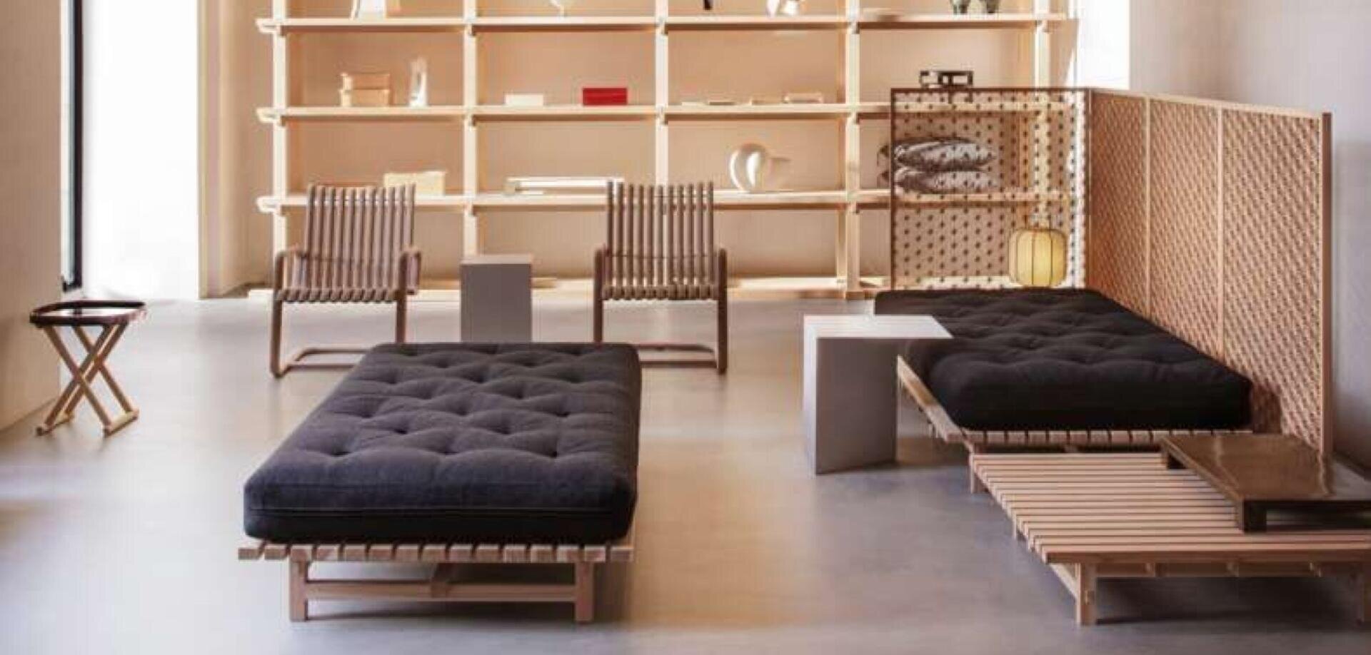 Minimalistisches Wohnzimmer mit Holzmöbeln, schwarzen gepolsterten Bänken, Lattenstühlen, einem niedrigen Tisch und einem hohen Regal mit Büchern und Dekorationsgegenständen vor einer neutral getönten Wand.