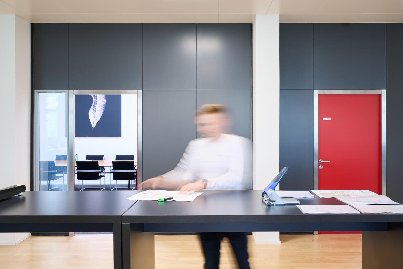 Eine Person in Bewegung steht an einem modernen Büro-Empfangstresen mit dunklen Paneelen, einer roten Tür und einem Konferenzraum, in dem im Hintergrund ein Gemälde zu sehen ist. Das Gesicht der Person ist aufgrund der Bewegung unscharf.