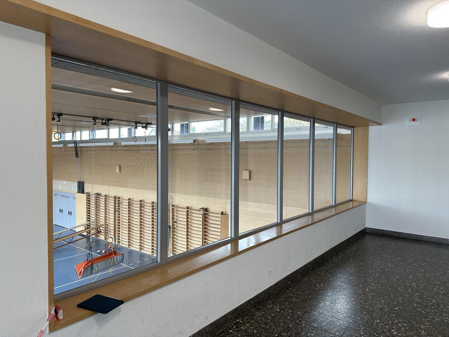 Blick durch große Innenfenster auf eine Turnhalle mit hölzerner Sprossenwand, blauem Boden und Basketballkorb, gesehen von einem Flur mit Terrazzoboden.