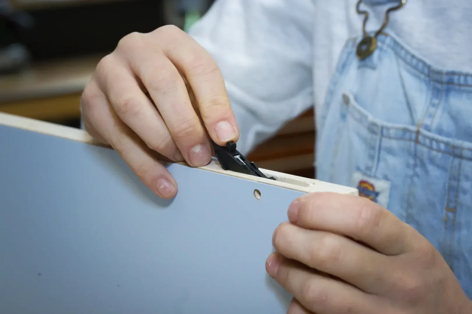 Eine Person, die einen hellblauen Overall und ein weißes Hemd trägt, benutzt ein schwarzes Universalmesser, um die Kante einer hellblauen laminierten Holzplatte zu schneiden.