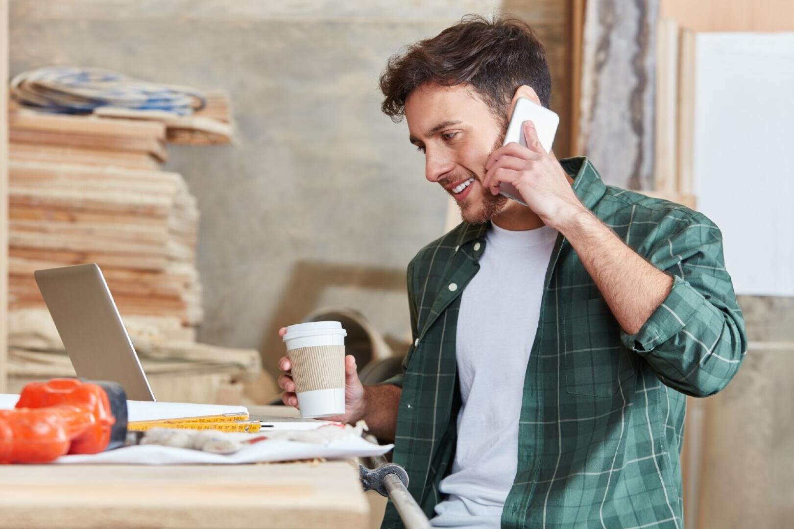 Ein Mann in einem grün karierten Hemd hält eine Kaffeetasse in der Hand und telefoniert, während er in einer Holzwerkstatt, umgeben von Werkzeugen und Holzbrettern, an einem Laptop arbeitet.