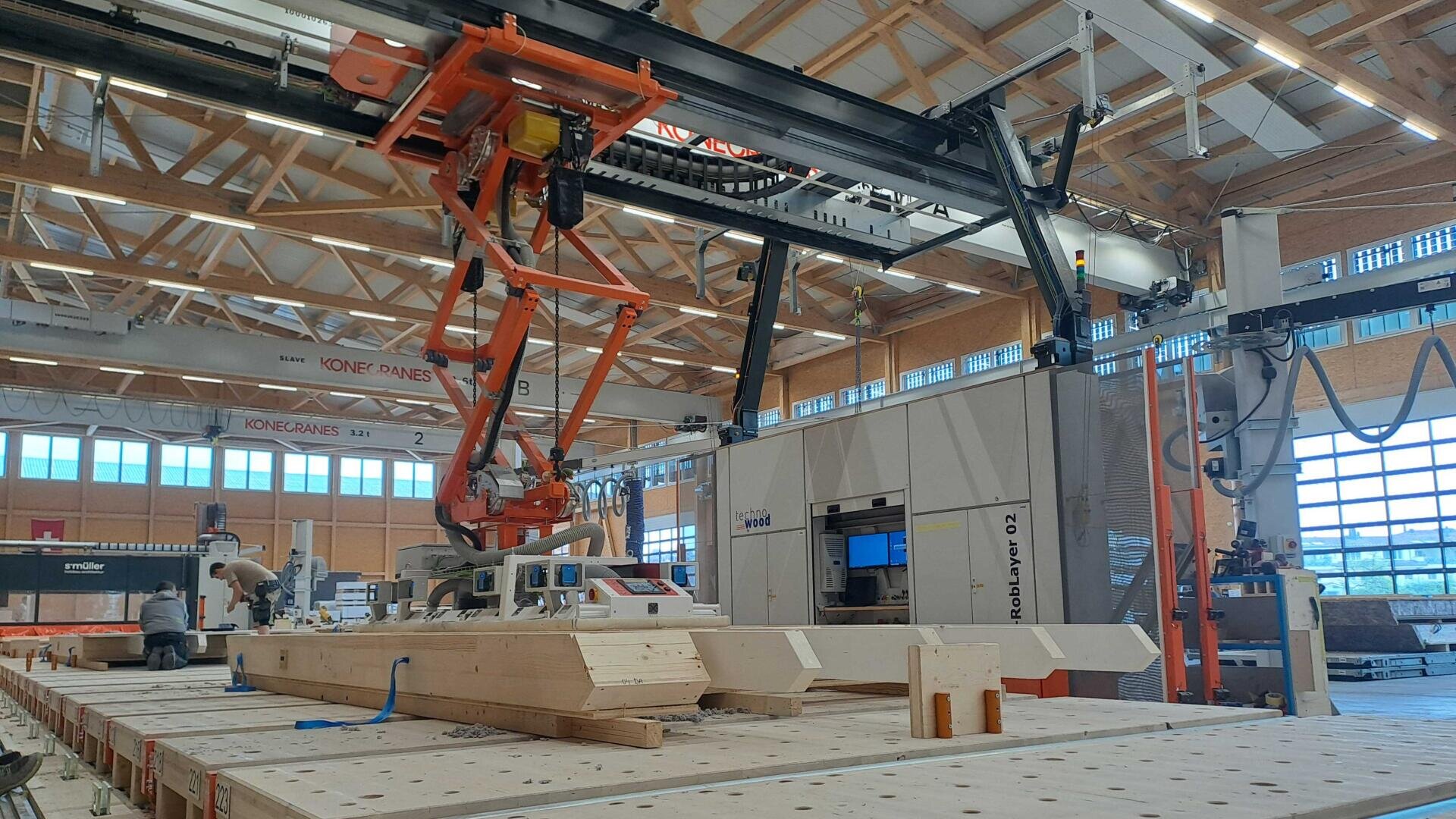 Un grand bras robotisé industriel assemble des poutres en bois à l'intérieur d'une usine spacieuse et moderne, avec de hauts plafonds et une lumière naturelle abondante provenant de grandes fenêtres. Diverses machines et équipements sont visibles à l'arrière-plan.
