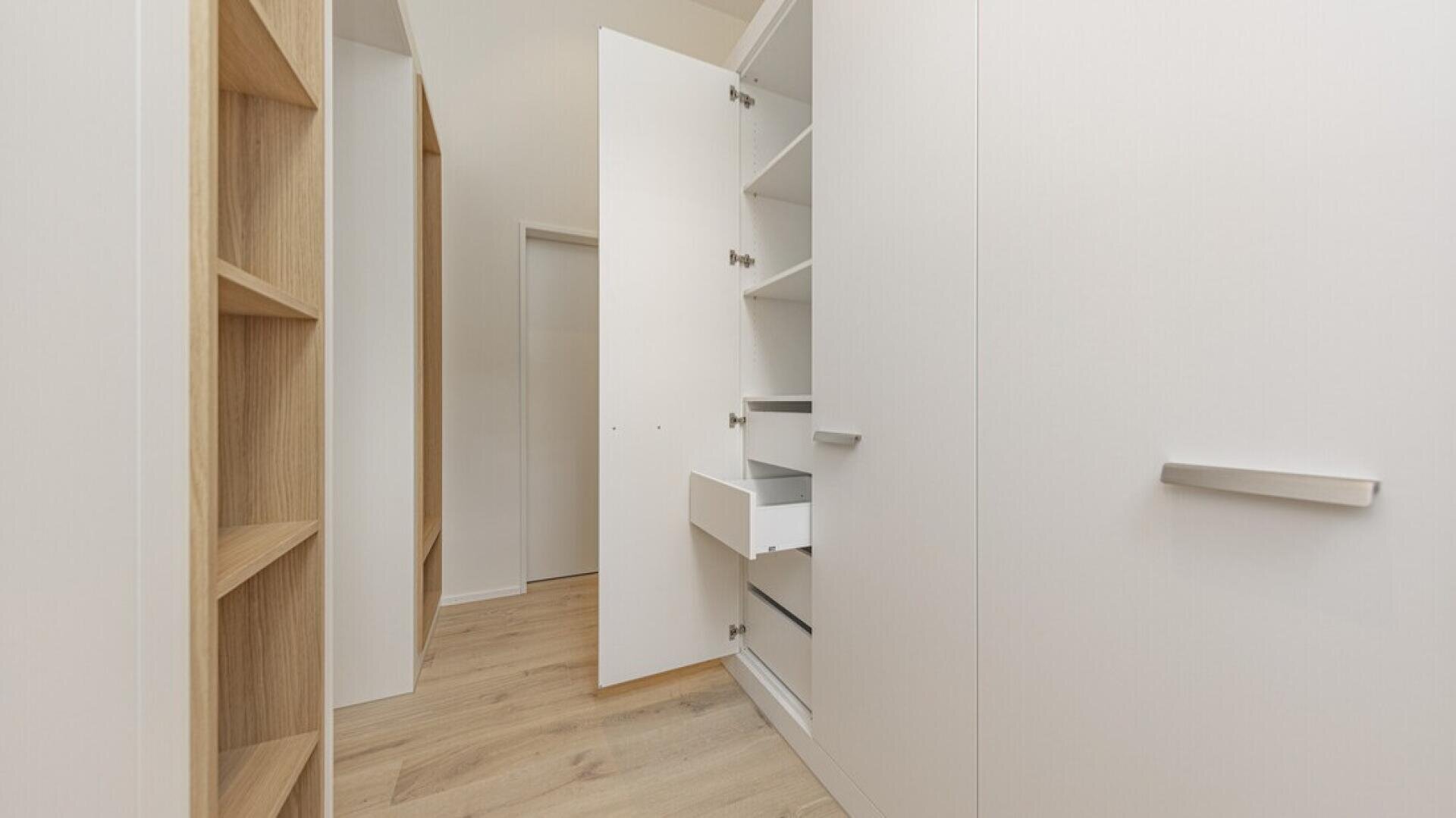 Un dressing moderne avec un sol en bois clair, des étagères en bois ouvertes sur la gauche et des armoires blanches avec des portes et des tiroirs ouverts sur la droite. L'espace est lumineux et minimaliste.