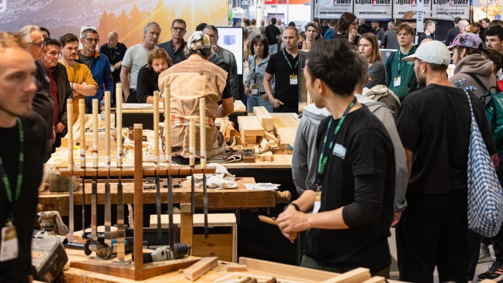 Eine Menschenmenge versammelt sich um eine Holzbearbeitungsvorführung auf einer belebten Veranstaltung und beobachtet eine Person, die mit Werkzeugen und Holz an einem Tisch arbeitet, wobei verschiedene Meißel und Geräte im Vordergrund zu sehen sind.