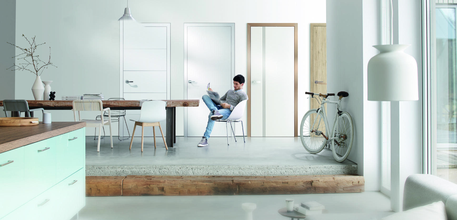 Ein Mann sitzt auf einem Stuhl und schaut auf sein Telefon in einem modernen, hellen Raum mit vier geschlossenen Türen, einem hölzernen Esstisch, weißen Stühlen, einem Fahrrad und einer minimalistischen Einrichtung. Sonnenlicht strömt durch große Fenster.