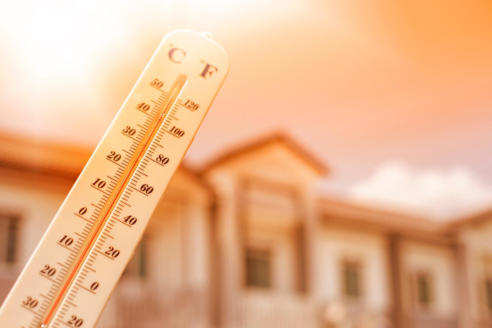 Ein Thermometer zeigt eine hohe Temperatur sowohl in Celsius als auch in Fahrenheit vor dem unscharfen Hintergrund eines Hauses unter hellen, sonnigen und warmen Bedingungen an.