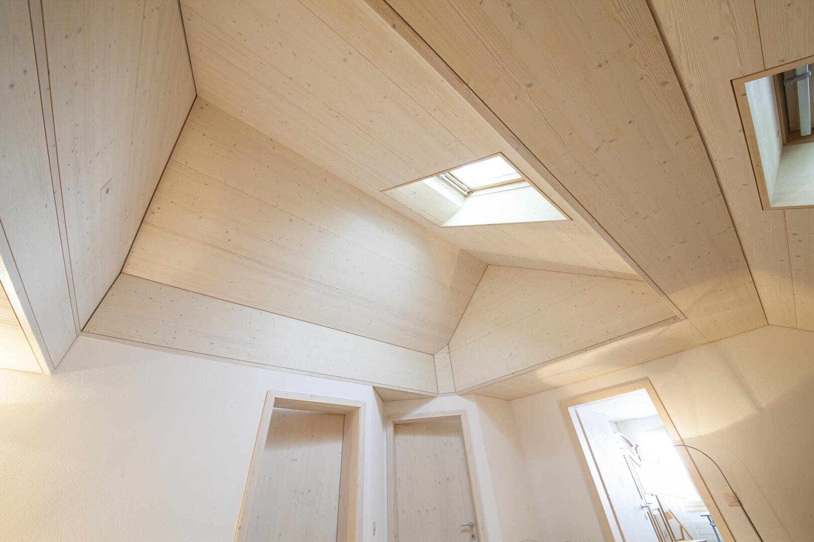 Vue en angle d'une pièce moderne avec des panneaux de plafond en bois clair, présentant des formes géométriques et une lucarne laissant entrer la lumière naturelle au-dessus de deux portes fermées.