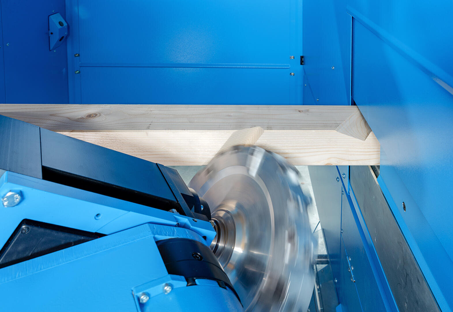 Eine Kreissäge mit blauem Gehäuse schneidet durch ein Stück helles Holz in einer Industriemaschine. Das Sägeblatt dreht sich schnell, wodurch ein Bewegungsunschärfe-Effekt entsteht.