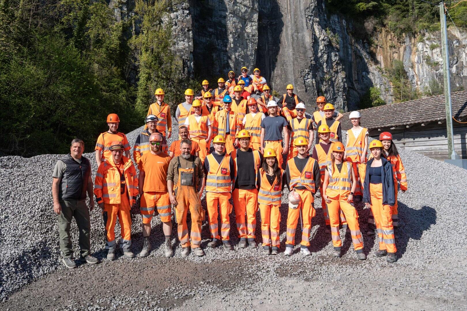 Un grand groupe de personnes portant des gilets de sécurité orange, des casques de protection et des équipements de travail posent ensemble sur du gravier devant une falaise rocheuse, avec des arbres et un bâtiment en bois à l'arrière-plan.