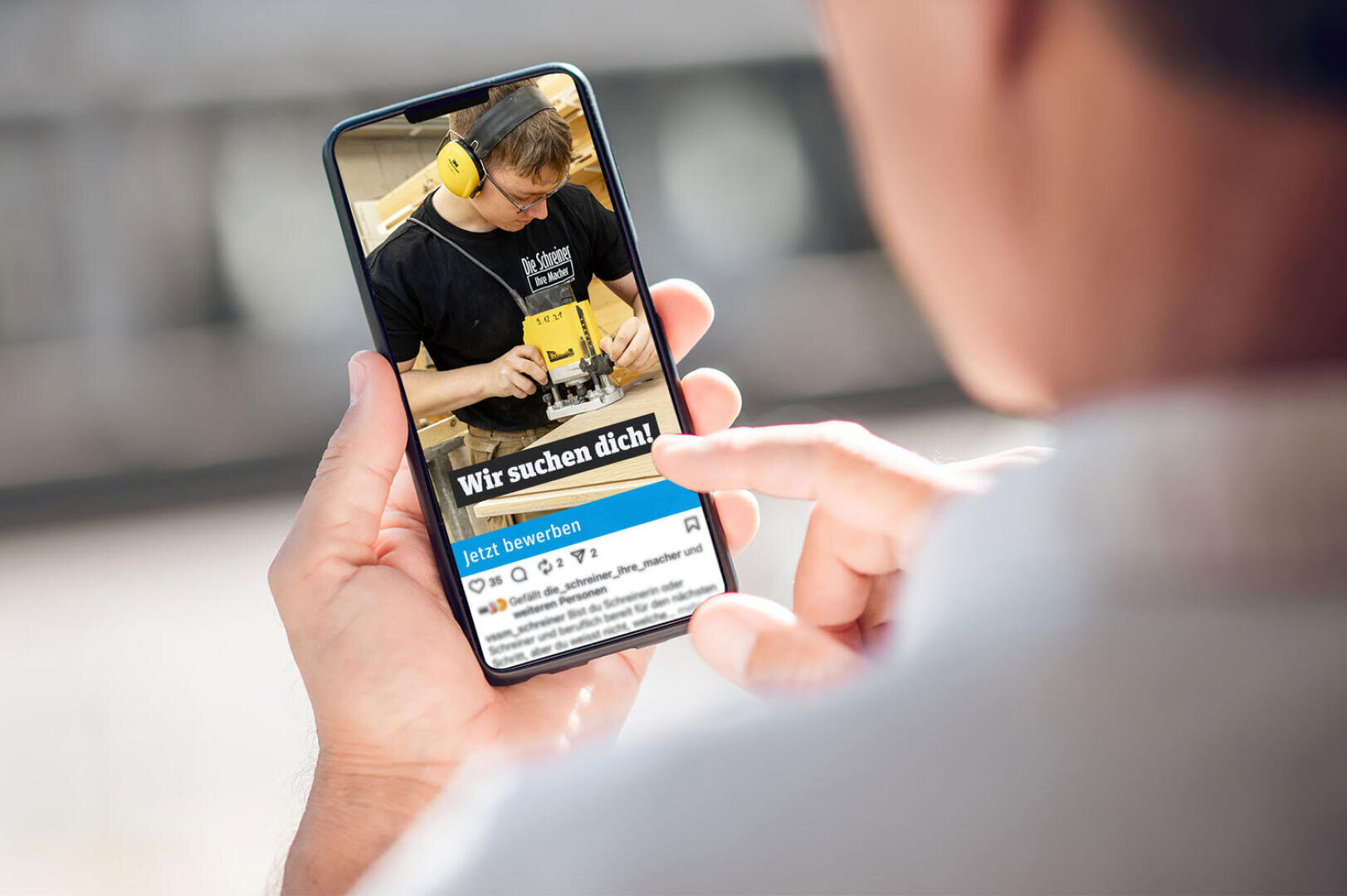 Eine Person hält ein Smartphone in der Hand und betrachtet eine Stellenanzeige, die einen Mann bei der Arbeit mit einem Holzbearbeitungswerkzeug zeigt. Die Überschrift der Anzeige lautet "Wir suchen dich!" ("Wir suchen dich!") und eine blaue Schaltfläche mit der Aufschrift "Jetzt bewerben".