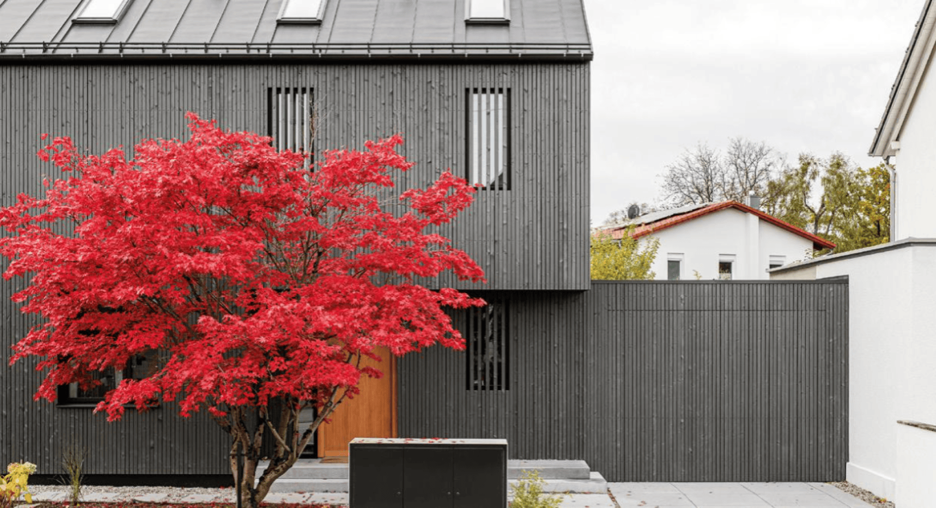 Ein modernes Haus mit dunkelgrauer, vertikaler Verkleidung hat schmale Fenster und eine Holztür. Im Vorgarten steht ein leuchtend rotblättriger Baum, der einen Kontrast zur minimalistischen Architektur bildet.