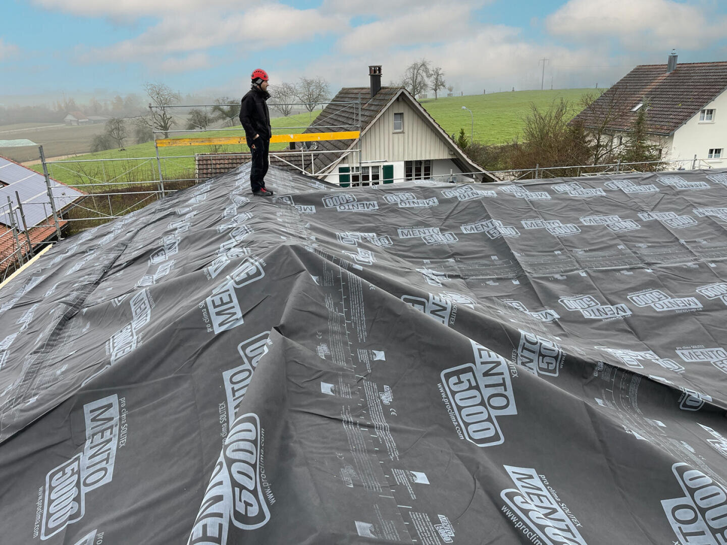 Eine Person mit Schutzausrüstung steht auf einem Hausdach, das mit schwarzen, wasserdichten Membranen abgedeckt ist. Im Hintergrund sind die benachbarten Häuser und eine Graslandschaft zu sehen.