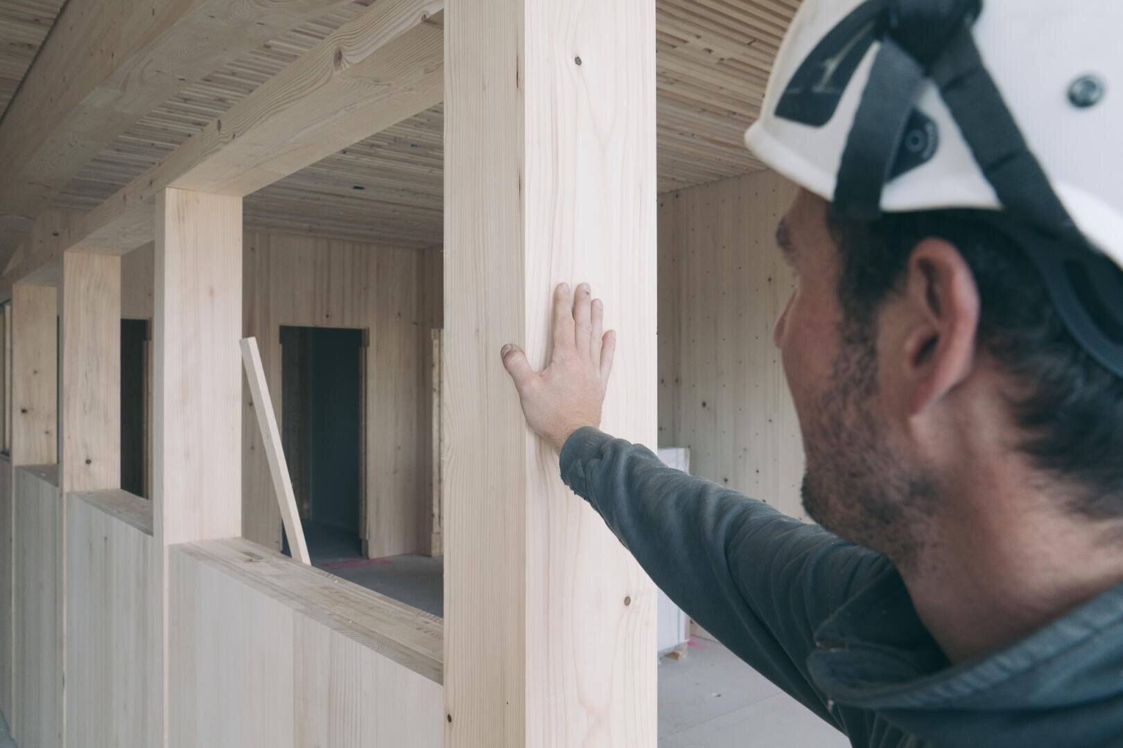 Eine Person, die einen weißen Schutzhelm trägt, berührt einen Holzpfeiler in einem im Bau befindlichen Gebäude mit freiliegenden Holzwänden und -balken.