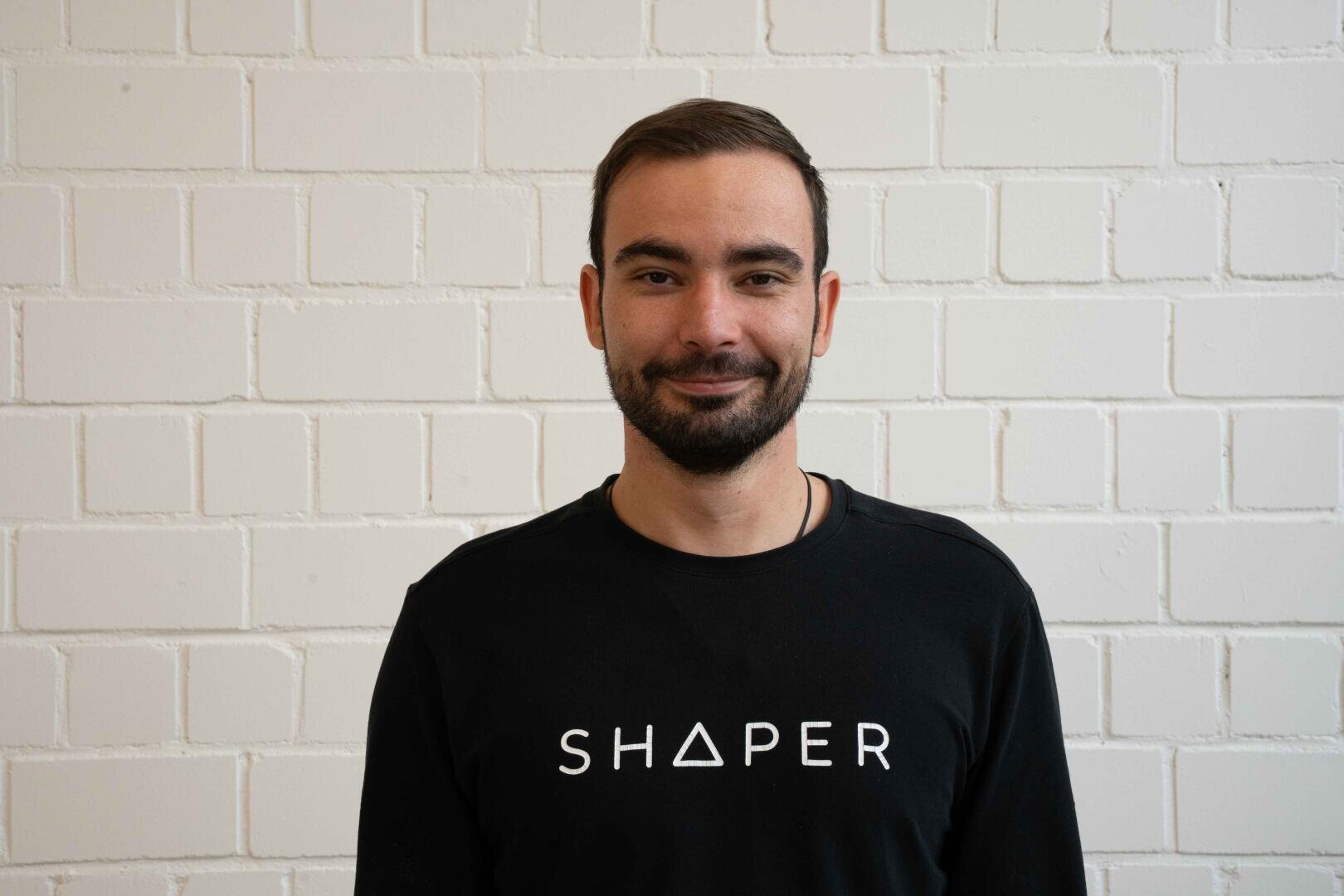 Ein Mann mit kurzen braunen Haaren und Bart steht vor einer weißen Backsteinmauer und trägt ein schwarzes Hemd, auf dem in weißen Buchstaben das Wort SHAPER steht.