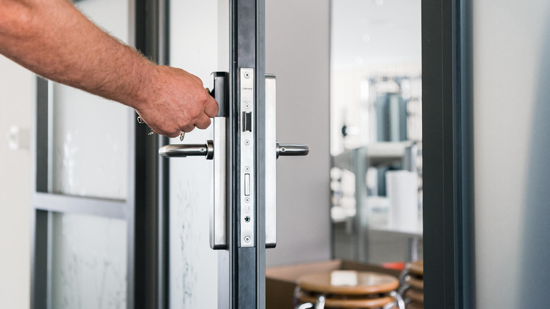 La main d'une personne tourne une clé dans la serrure d'une porte vitrée, l'ouvrant pour révéler un intérieur moderne et lumineux avec des chaises et des tables en arrière-plan.