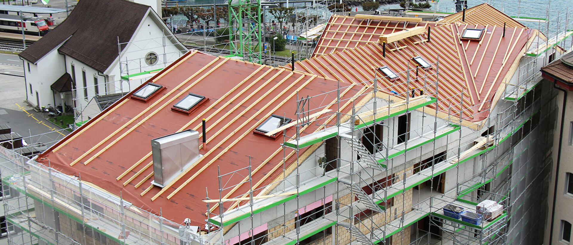 Ein im Bau befindliches Gebäude, umgeben von Gerüsten, dessen Dach teilweise fertiggestellt ist und auf dem bereits Holzbalken und Dachgauben angebracht sind. Auf der Baustelle sind Baumaterialien und Werkzeuge zu sehen.