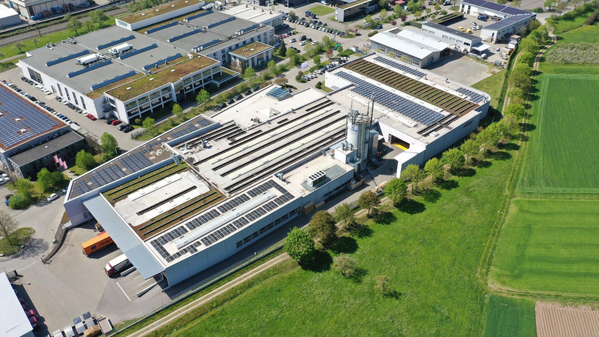 Luftaufnahme eines Industriekomplexes mit mehreren großen Gebäuden, von denen viele mit Solarzellen auf ihren Dächern ausgestattet sind, umgeben von Parkplätzen, Grünflächen und angrenzenden Feldern.