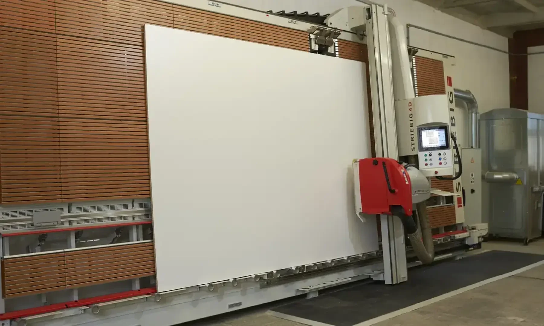Une grande machine à scier à panneaux verticale avec un panneau blanc, installée dans un hall industriel avec des murs en lattes de bois et des commandes de machine visibles à droite.