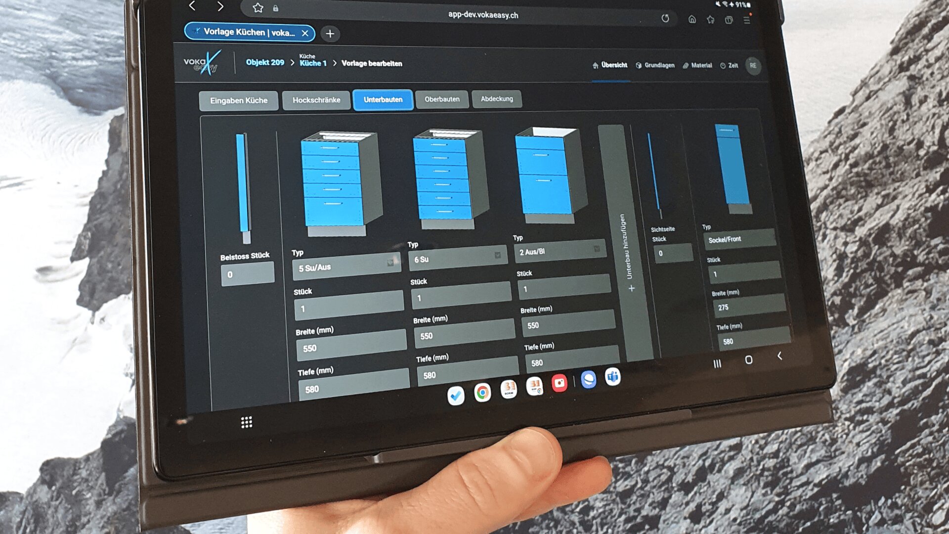 Eine Person hält ein Tablet, auf dem eine Planungssoftware-Schnittstelle mit blauen Küchenschränken, Abmessungen und Spezifikationen angezeigt wird. Der Hintergrund ist ein Graustufenbild von Felsen oder einer Gebirgslandschaft.