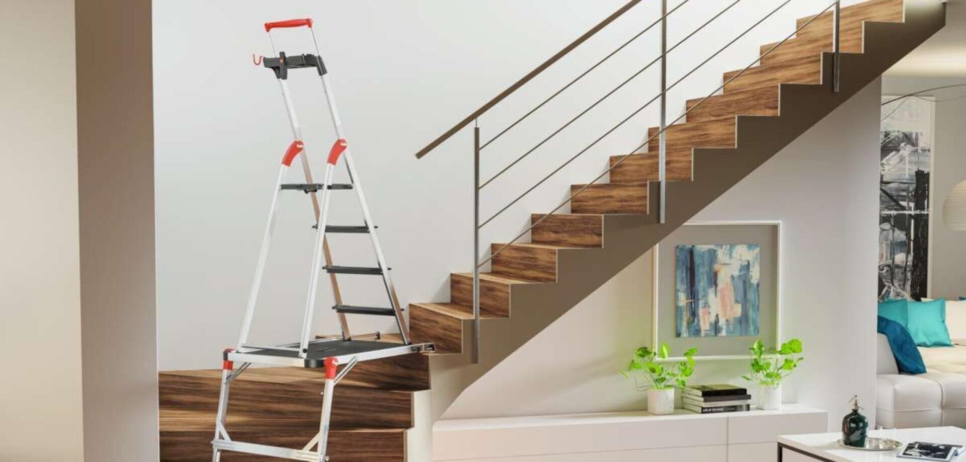 Eine klappbare Stufenleiter mit roten Griffen ist sicher auf einer Treppe angebracht und ermöglicht den Zugang zu höher gelegenen Bereichen. Das moderne Wohnzimmer hat eine Holztreppe, Kunstwerke, Pflanzen und eine Couch im Hintergrund.
