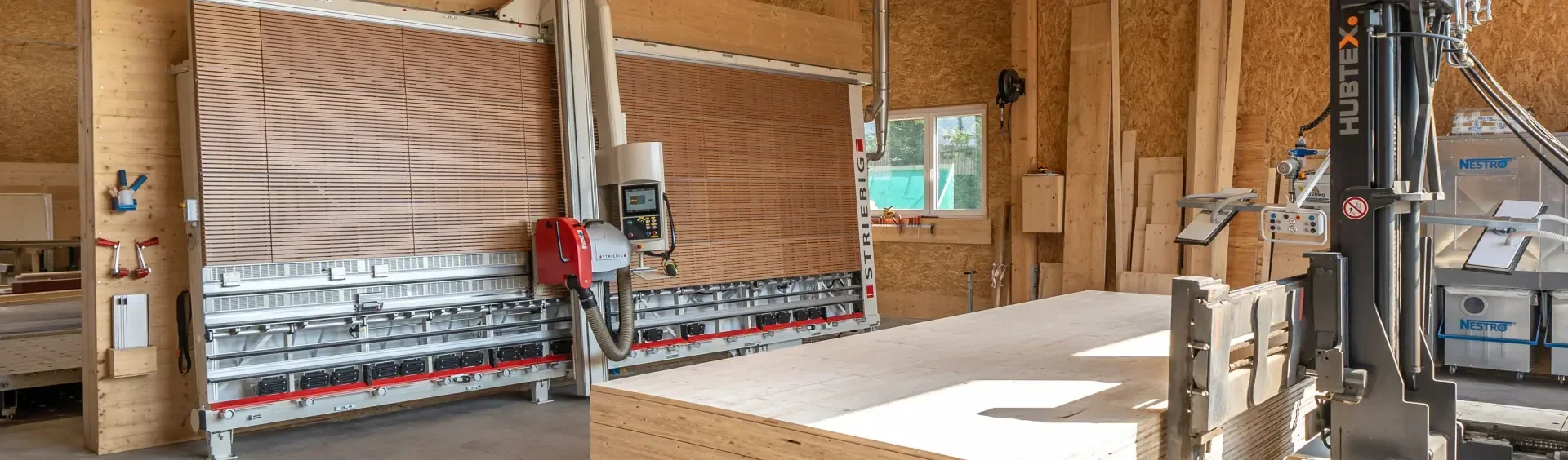 Un atelier de menuiserie avec de grandes scies à panneaux verticales, des murs lambrissés, des bois empilés et un chariot élévateur au premier plan. La lumière du soleil entre par une fenêtre et éclaire l'espace de travail organisé.