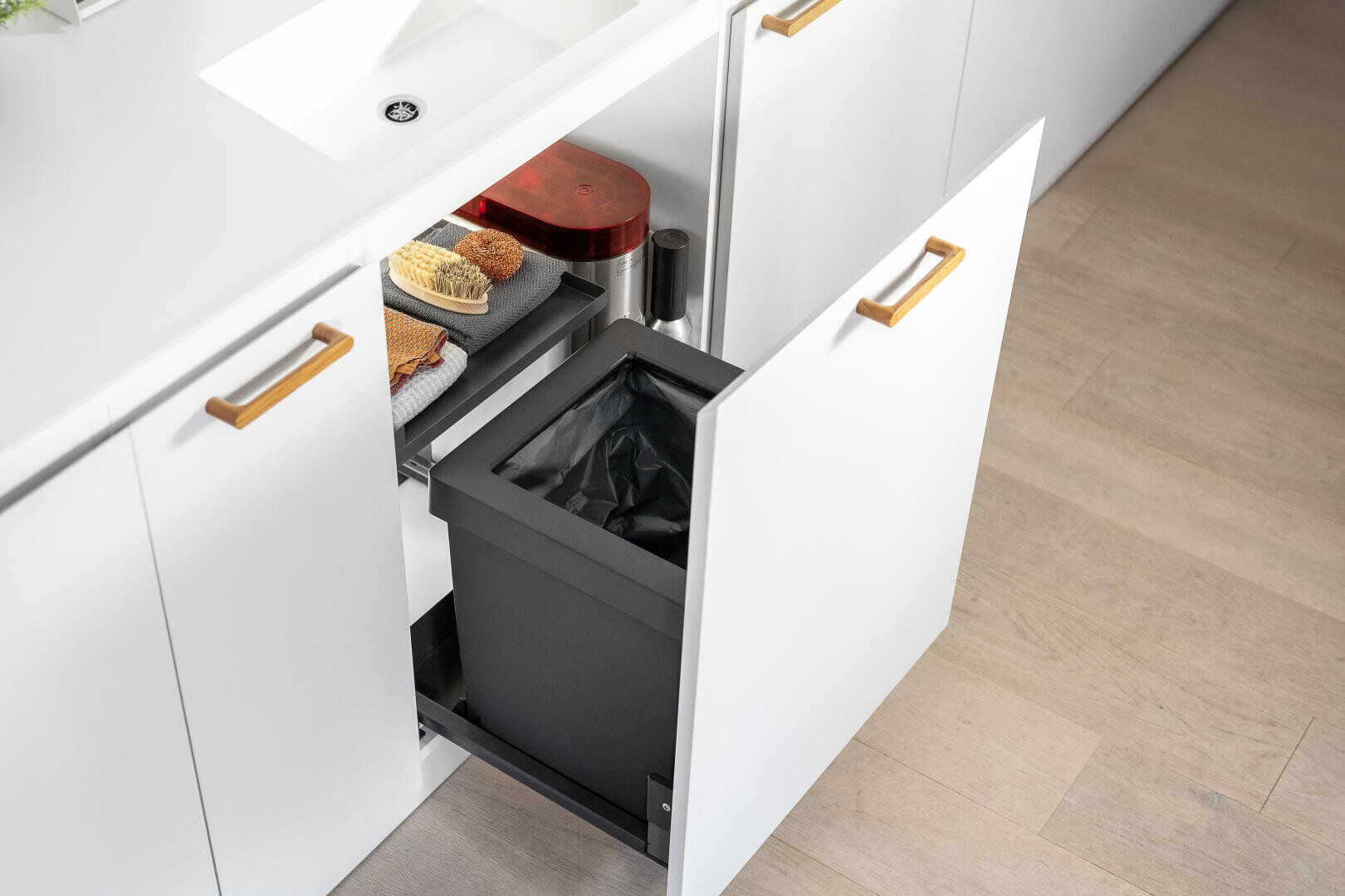 Une armoire de cuisine blanche ouverte montre une poubelle coulissante avec un bac noir sous une étagère fixe avec des éponges de nettoyage, une brosse et un récipient rouge. À côté du système de déchets se trouve un appareil de traitement de l'eau. L'armoire est placée sous un plan de travail blanc avec un évier.