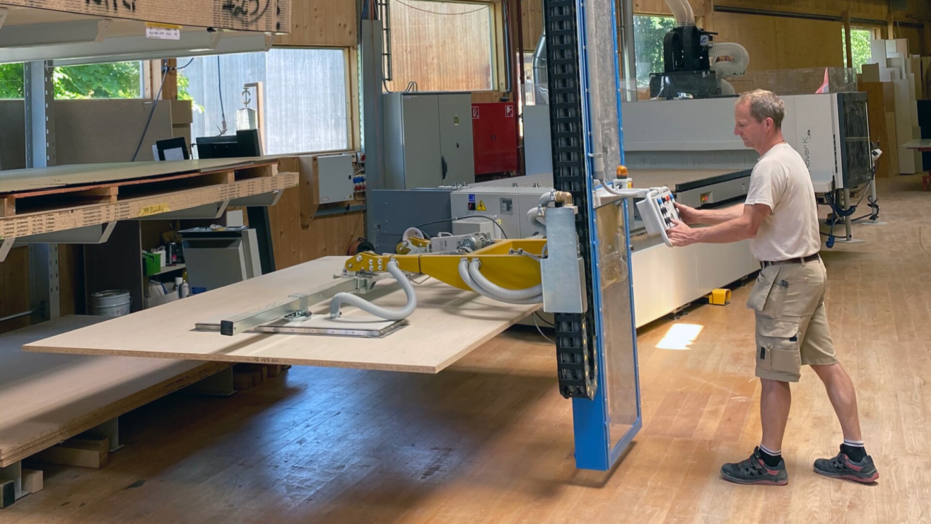 Une personne utilise une grande machine équipée d'un dispositif d'aspiration pour soulever et déplacer une feuille de bois dans un atelier spacieux et bien éclairé, rempli d'équipements de travail du bois.